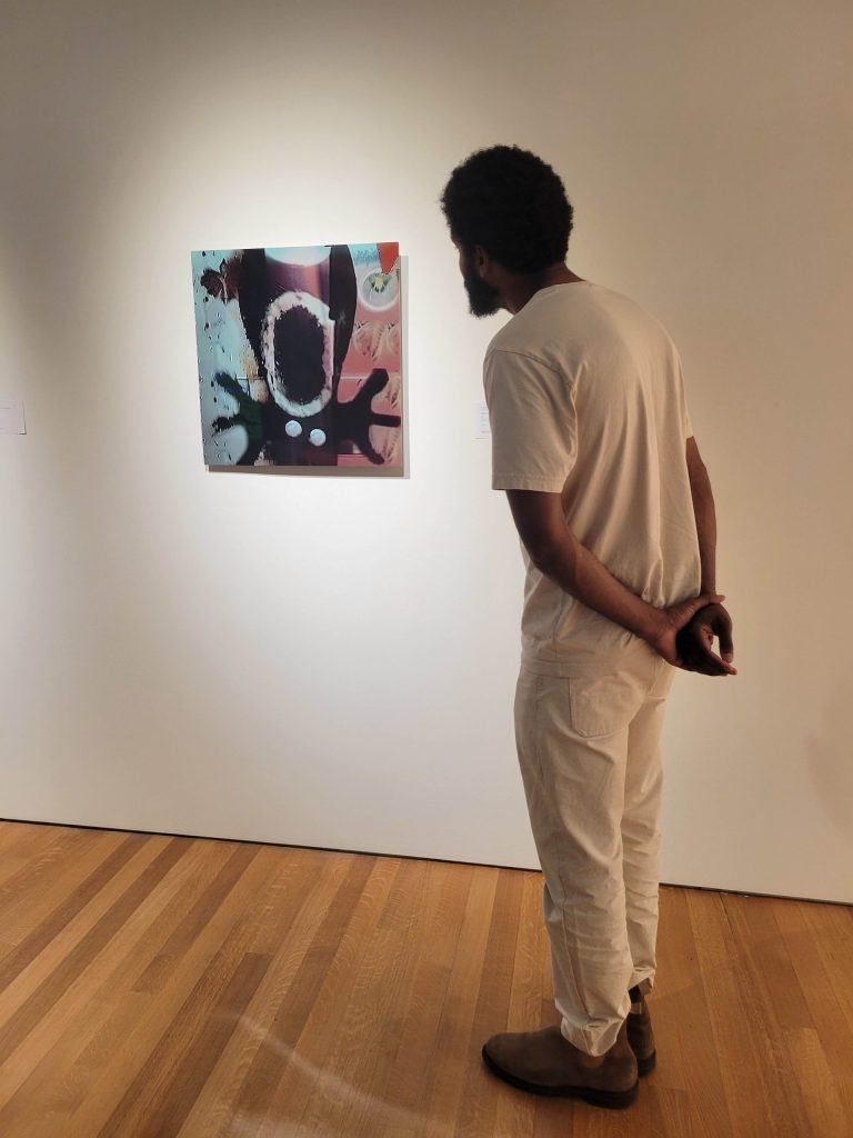 A gallery visitor in a cream T-shirt and trousers stands with his hands clasped behind his back, leaning in toward Mickey Finn by Will Martin, a square aluminum-plate print of a distorted cartoon face, hung on a white wall at Sotheby's New York.